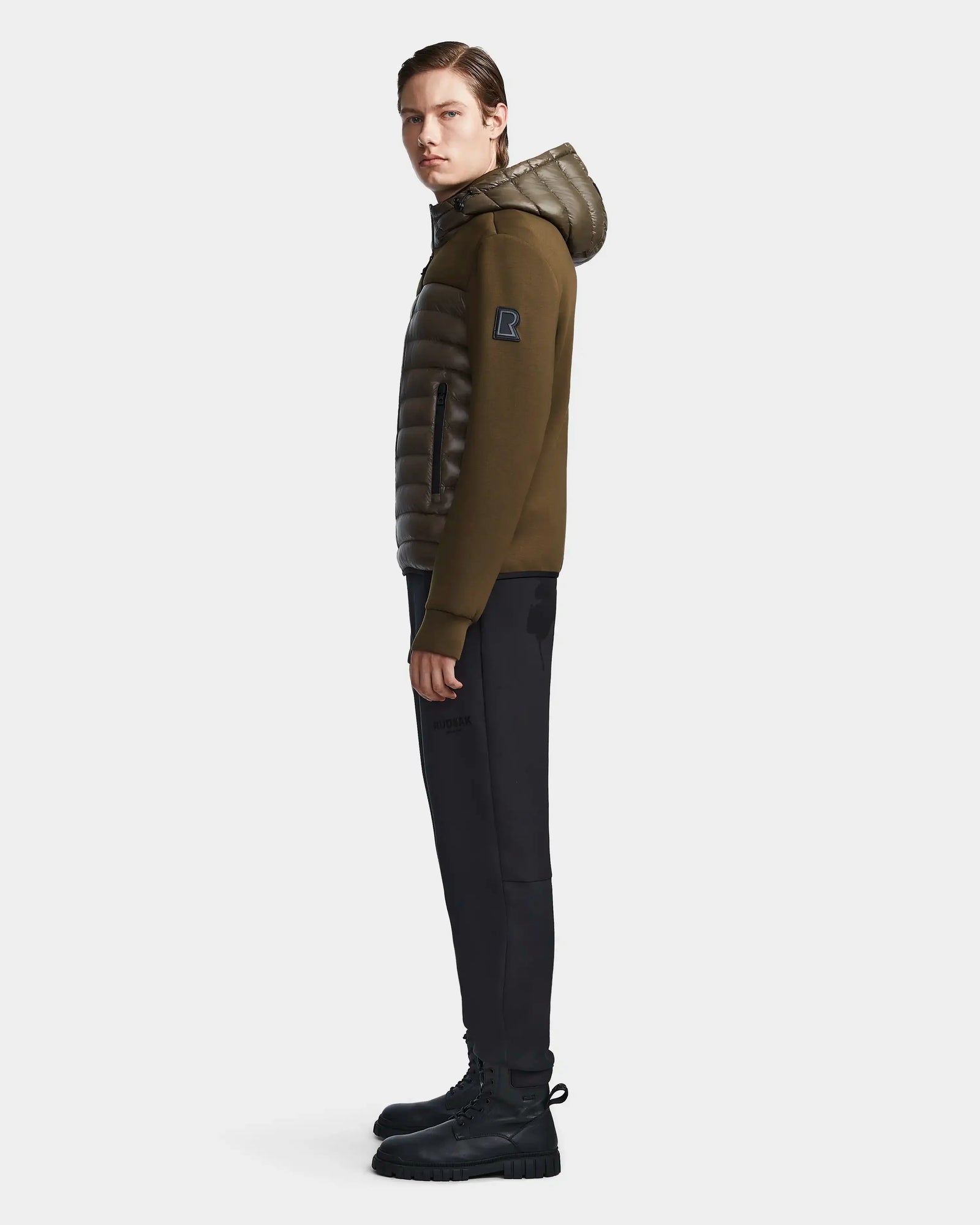 A man stands in profile against a white background wearing the Rudsak DURIN S - M123518 Light Olive mens hooded jacket with lightweight down insulation, black sleeves, waterproof zippers, black pants, and boots.