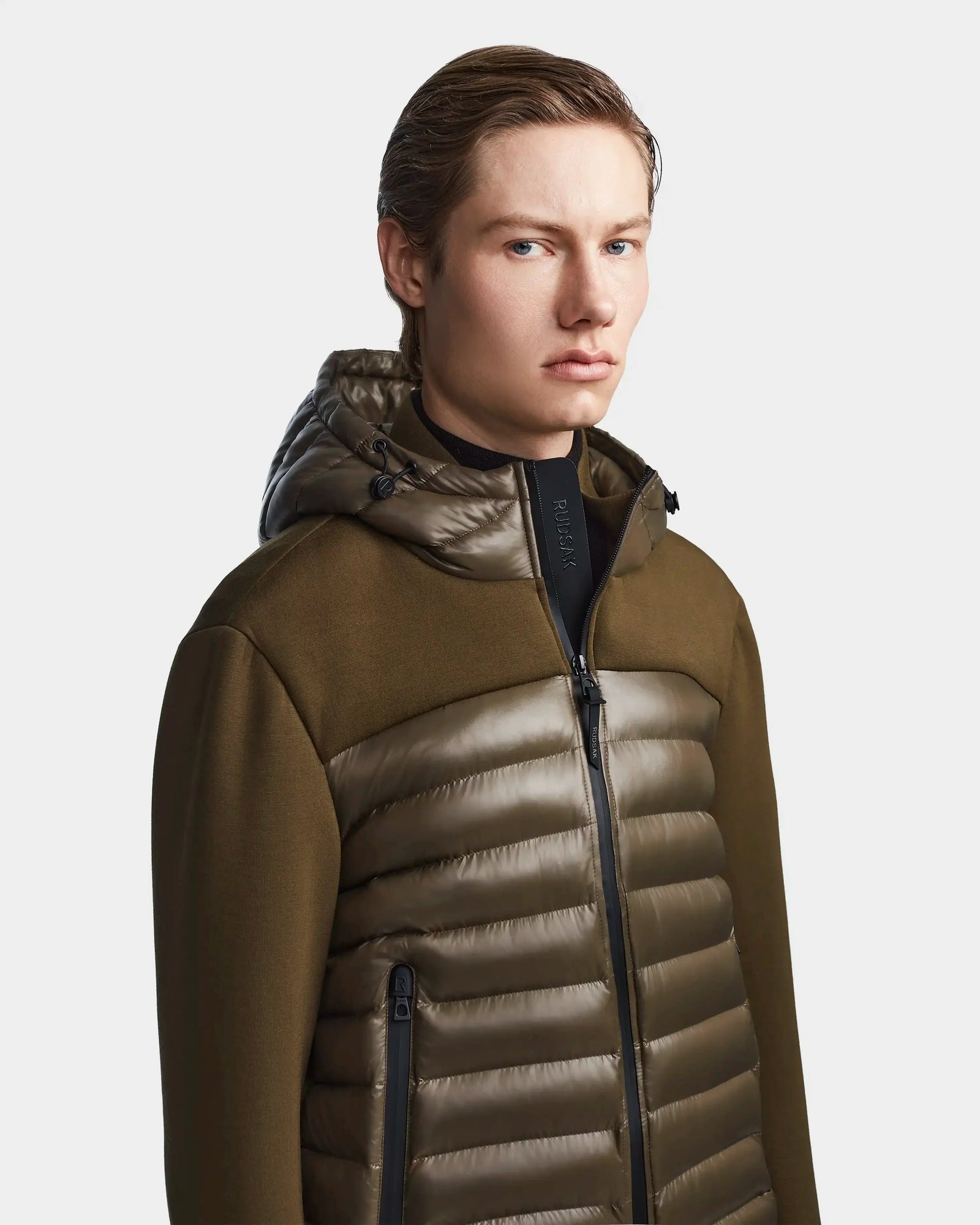 A young man with light skin and straight, light brown hair wears the Rudsak DURIN S - M123518 Light Olive mens hooded jacket, featuring waterproof zippers and lightweight down insulation, against a plain light background.