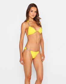 Model wearing the 'Raja Skimpy Bottom - Sunshine' bikini, showcasing a vibrant yellow design with dazzling crystal accents on the sides. Styled with a matching triangle top, she poses confidently against a white wooden wall.
