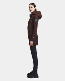 A woman in profile models the RUDSAK MERINA - 8122546 CHOCOLATE long quilted down jacket with a hood, styled with black leggings and chunky black boots, set against a plain light background.