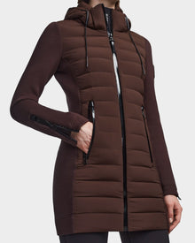 Wearing the RUDSAK MERINA - 8122546 CHOCOLATE quilted down jacket, a person showcases its long brown design with a hood, black zipper details, zippered pockets and cuffs, and fitted sleeves—perfect as a transitional piece with one hand in a front pocket.