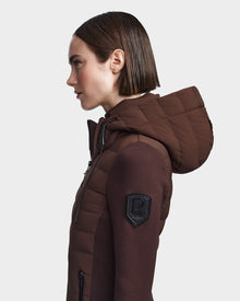 A woman with short brown hair models the RUDSAK MERINA - 8122546 CHOCOLATE hooded quilted down jacket, featuring an “R” patch on the sleeve, shot in profile against a white background. This jacket is perfect for any season.