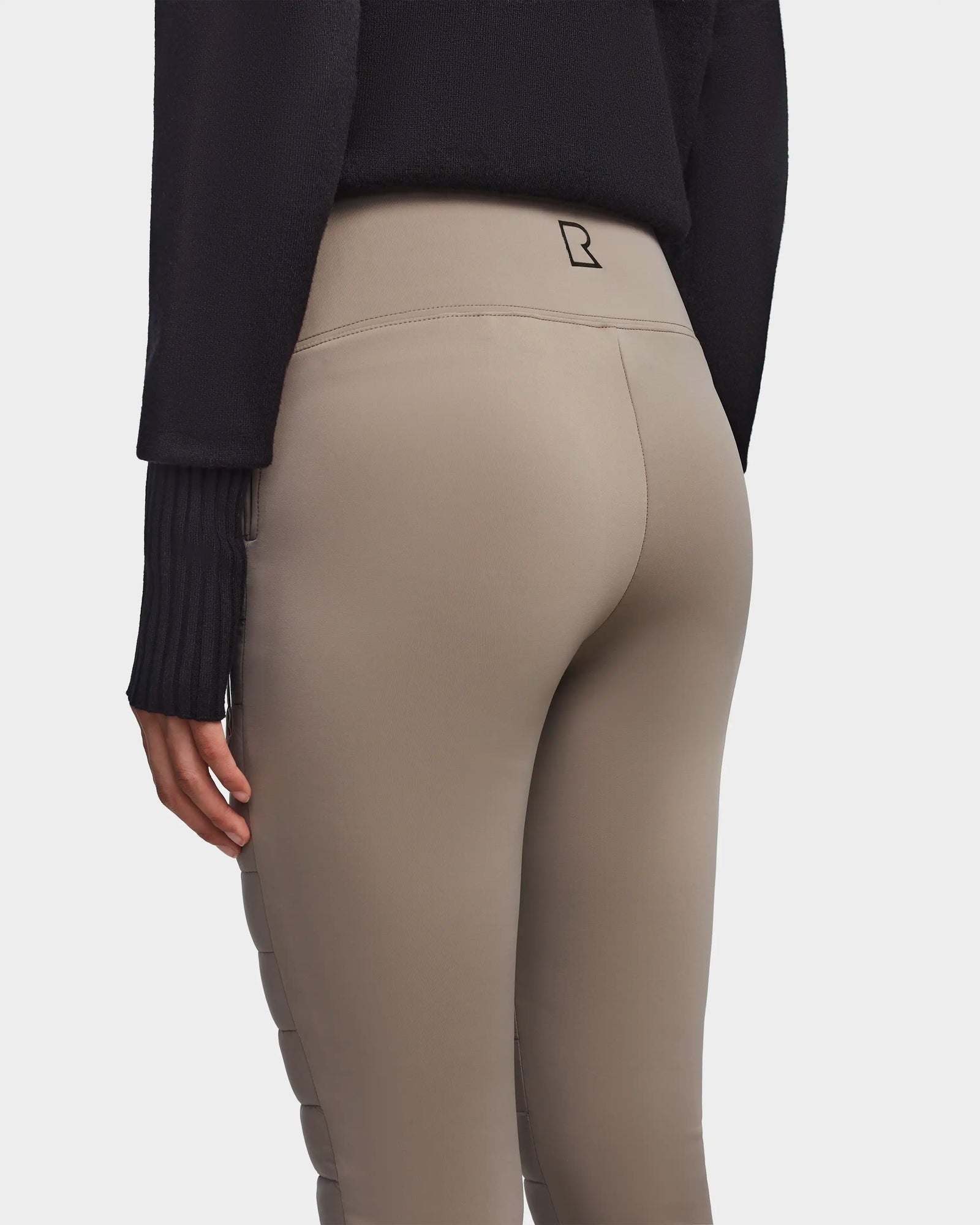 A person is shown from the back wearing Rudsak’s ARIL M - 8522514 Cement beige fitted sustainable winter pants with a small black logo at the waistband and a black long-sleeve sweater, their left arm relaxed at their side.