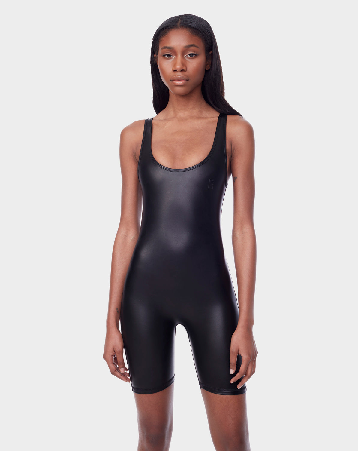 A woman with long, straight hair wears the Rudsak AVERI - 8523015 BLACK sleeveless fitted faux leather romper with a scoop neckline, standing against a plain light gray background.