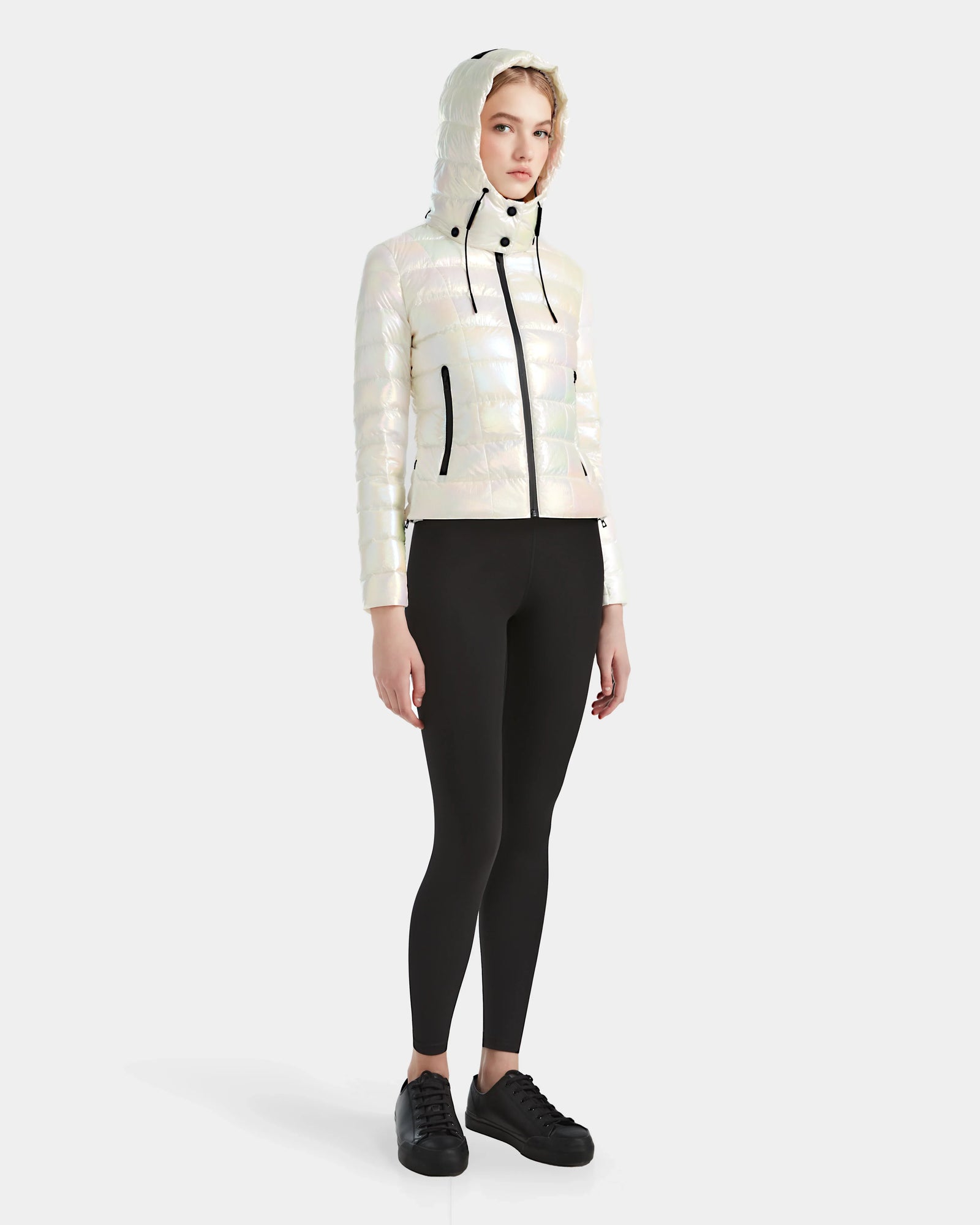 A person faces forward in a Rudsak MELISSA - 8124031 Iridescent Eggshell womens lightweight down puffer jacket with a hood, paired with black leggings and black sneakers, set against a plain white background.