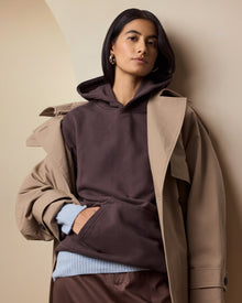 Person wearing a brown hoodie and beige coat against a beige wall