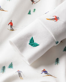 Close-up of Petite Plumes Babys Pima Romper in Apres Ski, showcasing a small embroidered green leaf on soft white Pima cotton fabric. The pattern includes skiers in vibrant red and blue attire with green pine trees. Leaf emblem is the focus.
