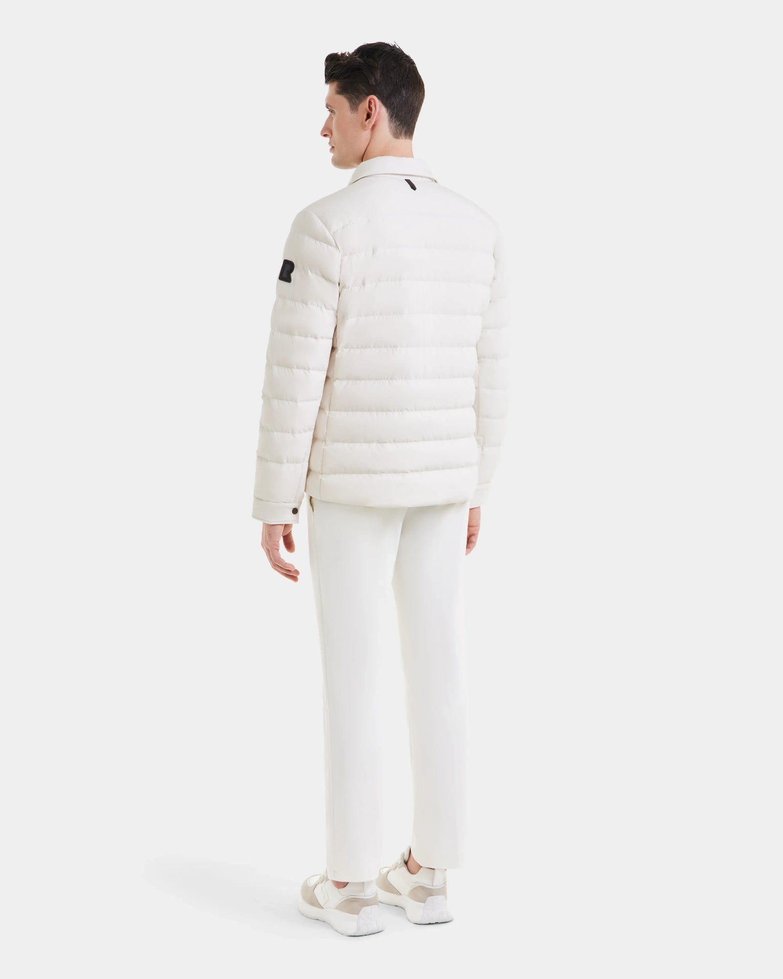 A man stands facing away, wearing the Rudsak AZIEL - M124521 Vanilla duck down jacket with white pants and sneakers against a plain white background.