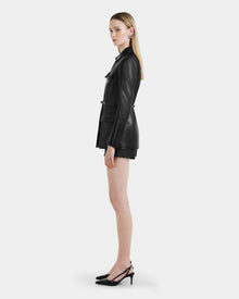 A woman with straight blonde hair stands in profile wearing the Rudsak DANICA - 8125025 Black leather jacket, paired with black shorts and slingback heels, against a plain light background.