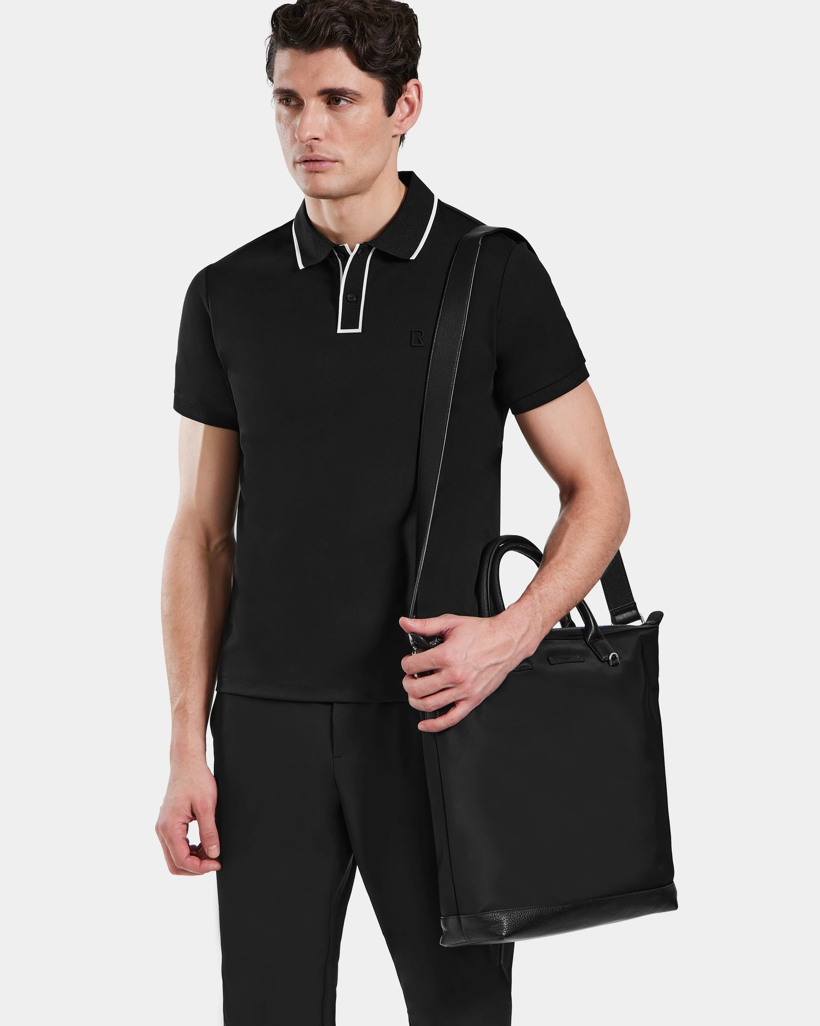 A man in a black polo with white trim and black pants stands against a plain background, holding the Rudsak DEXTER - 9324513 Black unisex tote by its handles and shoulder strap.