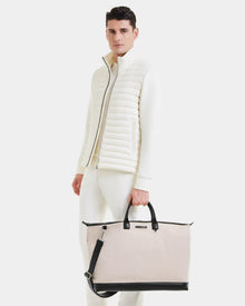 A man in an all-white outfit and quilted vest holds the Rudsak DUBLIN - 9324514 Vanilla unisex weekender tote, featuring handles and a shoulder strap, standing against a plain white background.