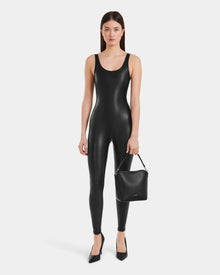 A woman stands against a plain background wearing the LAWREN - 8523047 Black sleeveless vegan leather bodysuit by Rudsak, paired with black pointed-toe heels and holding a black handbag in her left hand.