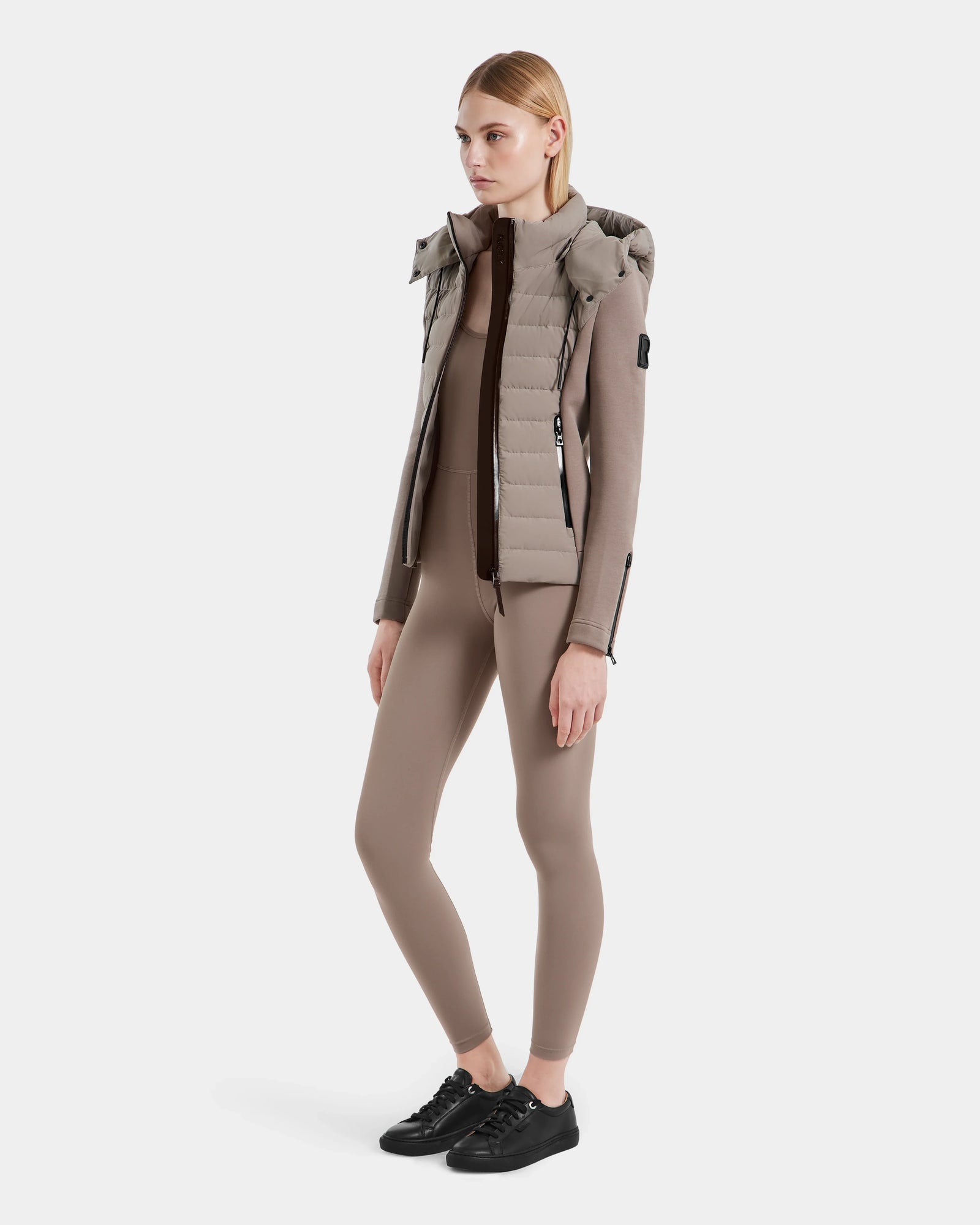 A woman faces slightly left, wearing the Rudsak MABS - 8124825 Cement lightweight short jacket with a hood, paired with a matching fitted top, leggings, and black sneakers against a plain white background.