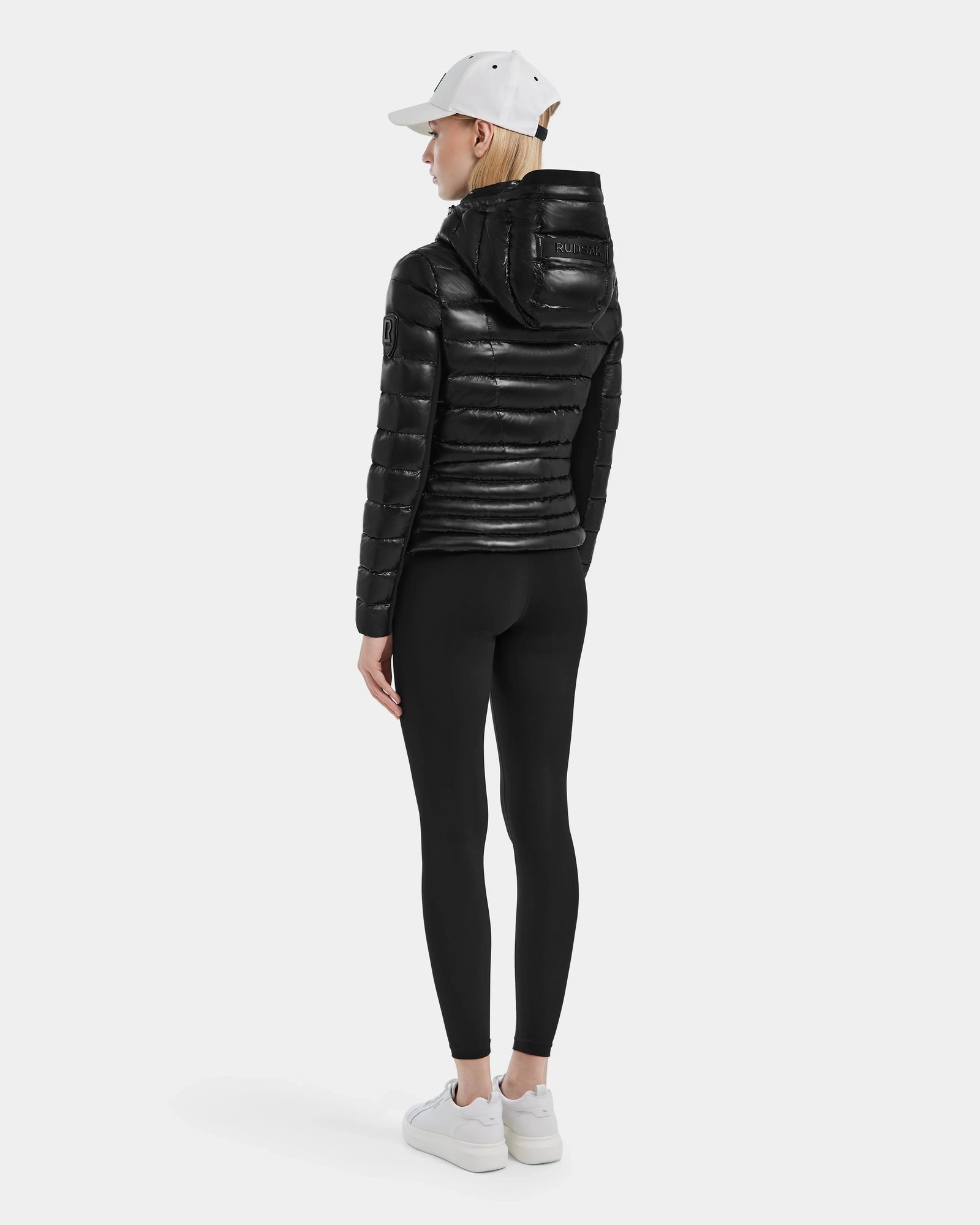 A person stands facing away in the MELISSA - 8121000 BLACK women’s puffer jacket, made from RDS certified down, paired with black leggings, white sneakers, and a white baseball cap against a plain light gray background.