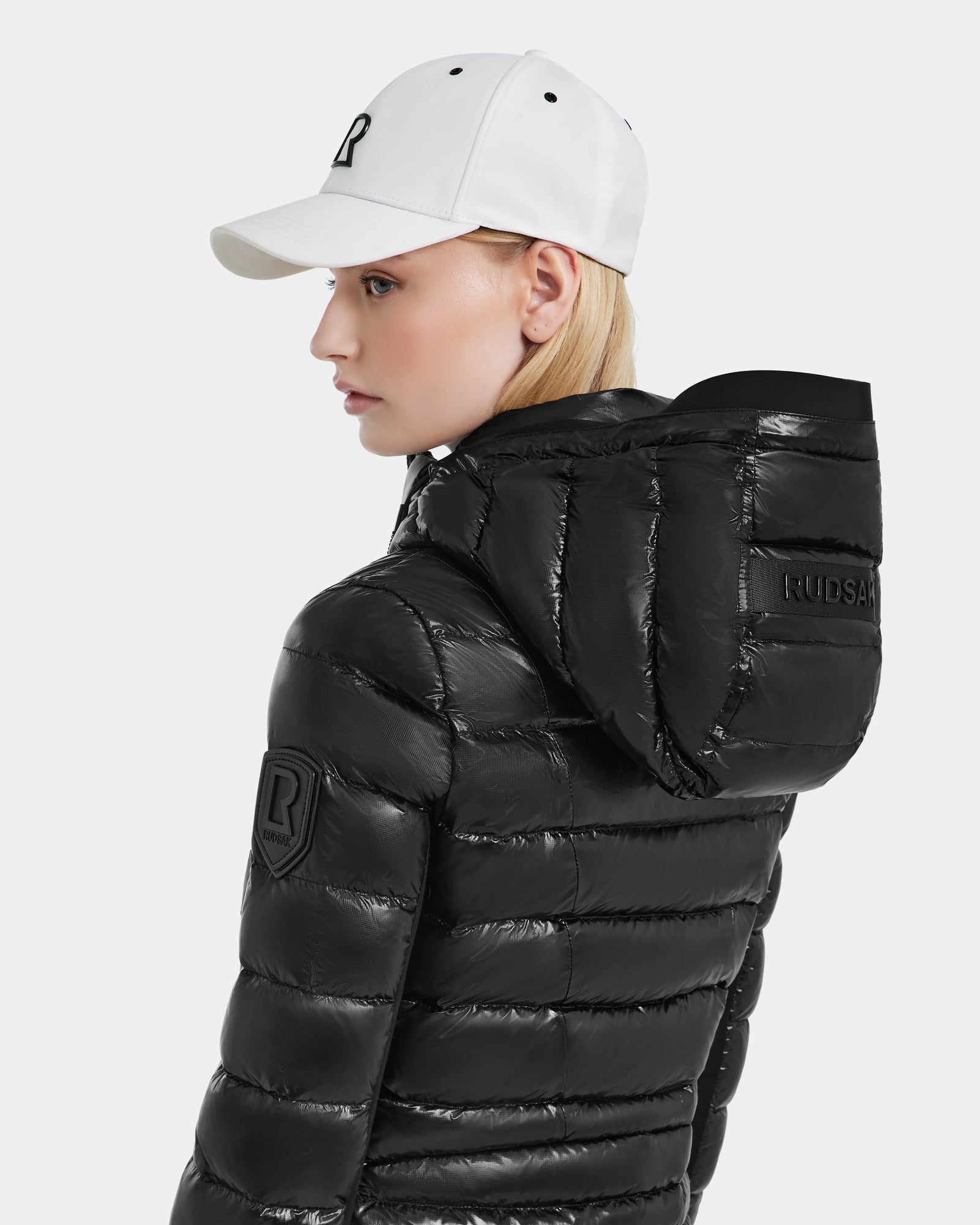 A woman wearing the MELISSA - 8121000 BLACK shiny black puffer jacket with a hood and a white baseball cap is shown in profile against a plain white background. The jacket features RDS certified down for warmth and responsible style.