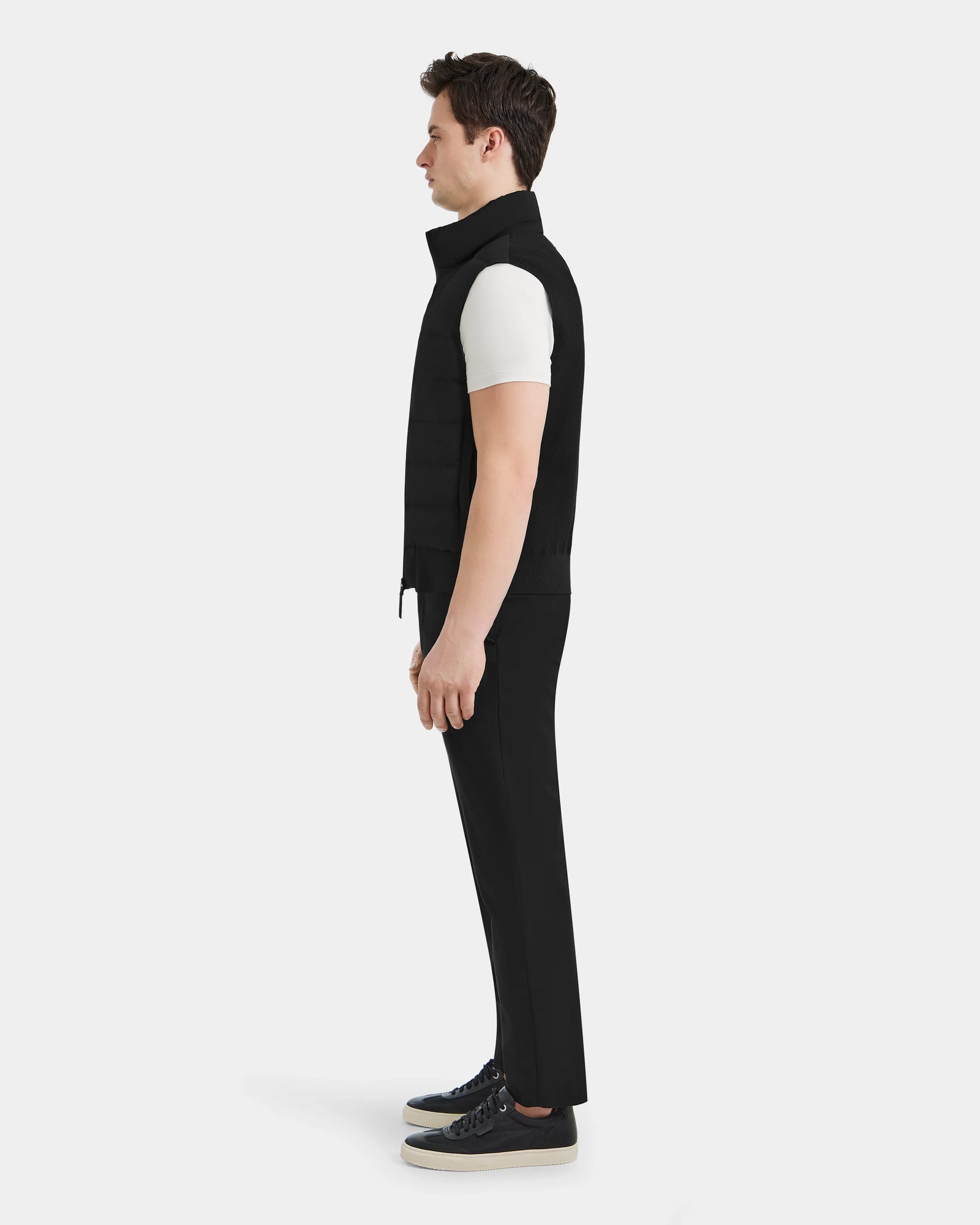 A man stands in profile against a white background, wearing the Rudsak MERLO - 6124017 Black men’s vest over a white short-sleeve shirt, with black pants and black sneakers.