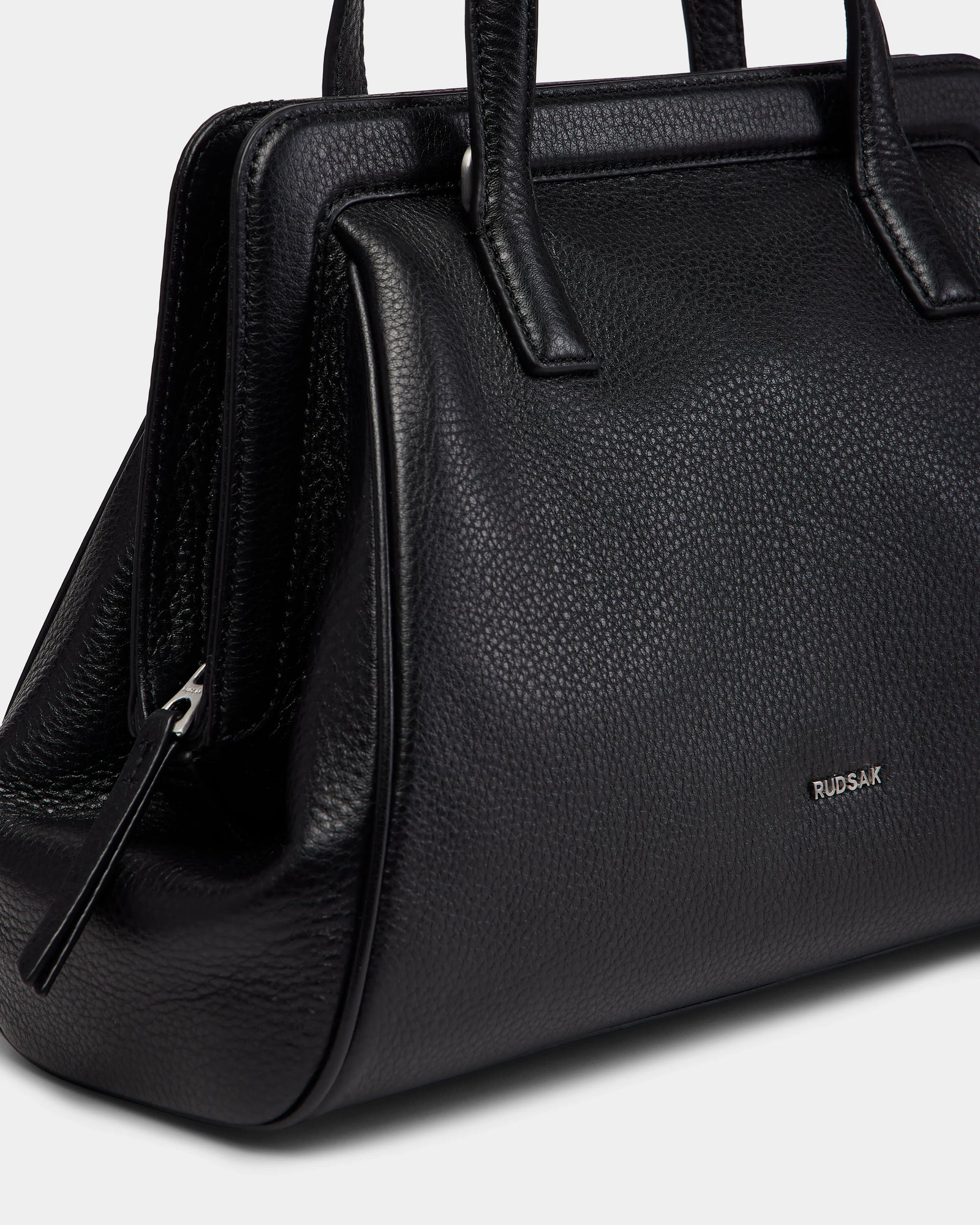 Close-up of the TAULLA - 8325002 Black women’s pebble leather satchel by Rudsak, featuring two handles, a spacious interior, a silver zipper, and the brand name in small silver letters on the side.