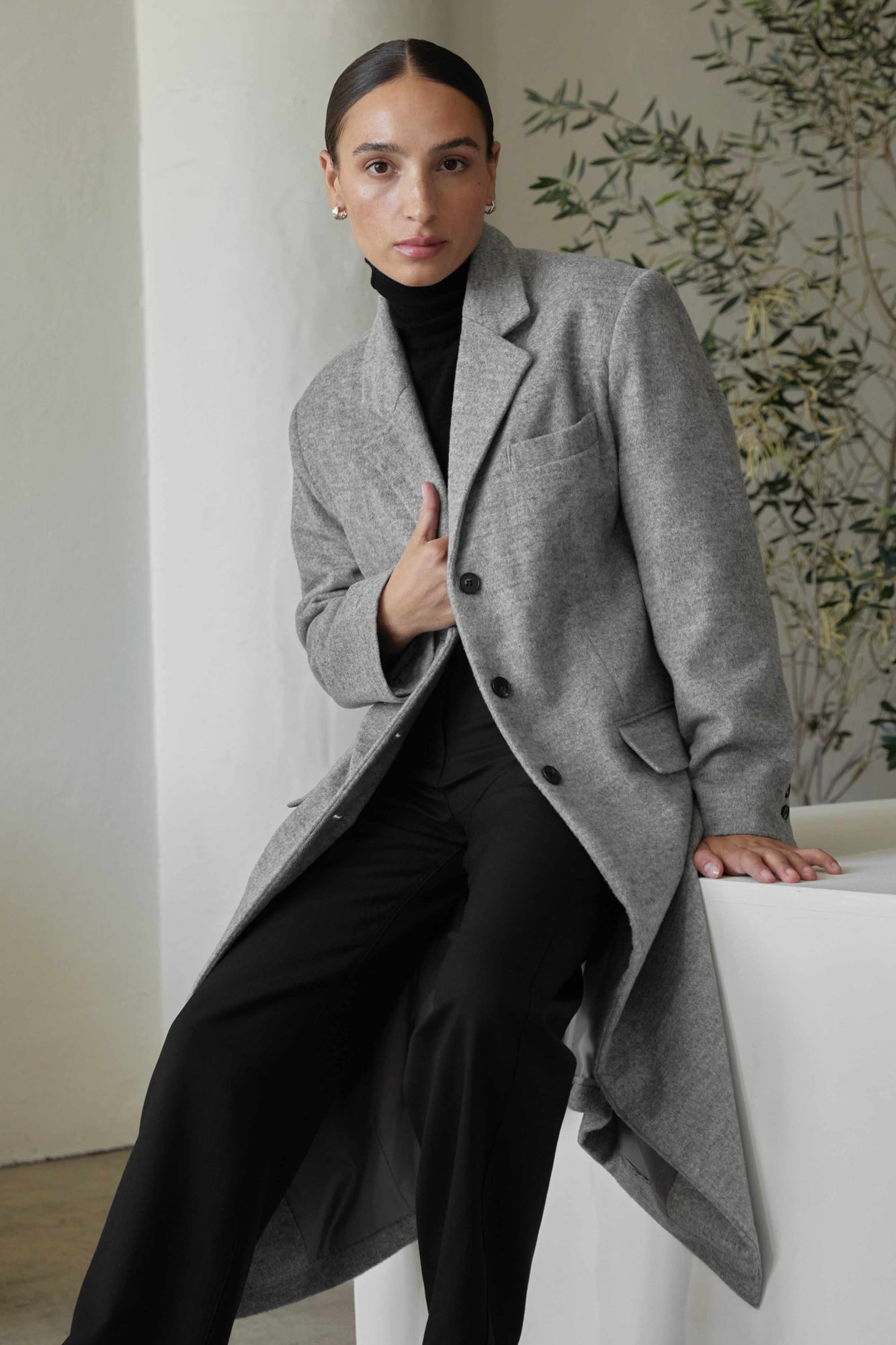 Woman wearing a gray coat over a black outfit, sitting on a white chair.