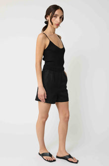 Woman wearing a black tank top and black linen shorts against a light grey background