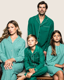A family of four poses in coordinated sleepwear against a plain background. The adults wear solid and checkered patterns, while the children don Petite Plumes Kids Flannel Pajama Set in Forest Green. Their expressions are serious as they sit closely together.