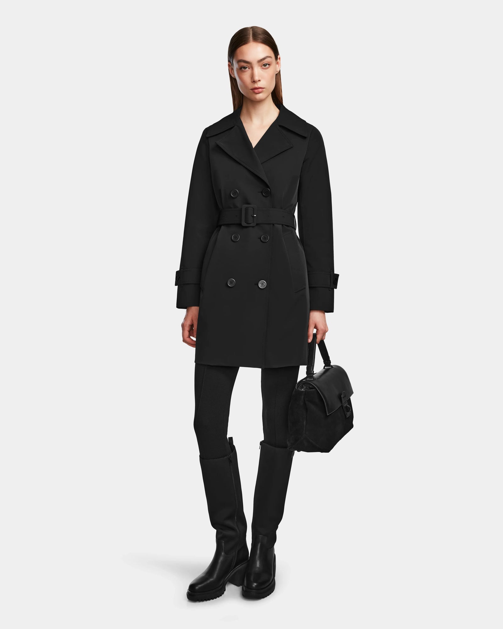 A woman wearing the Rudsak KIMBERLY - 8124801 Black belted trench coat, black leggings, black knee-high boots, and holding a black handbag stands against a plain light background.