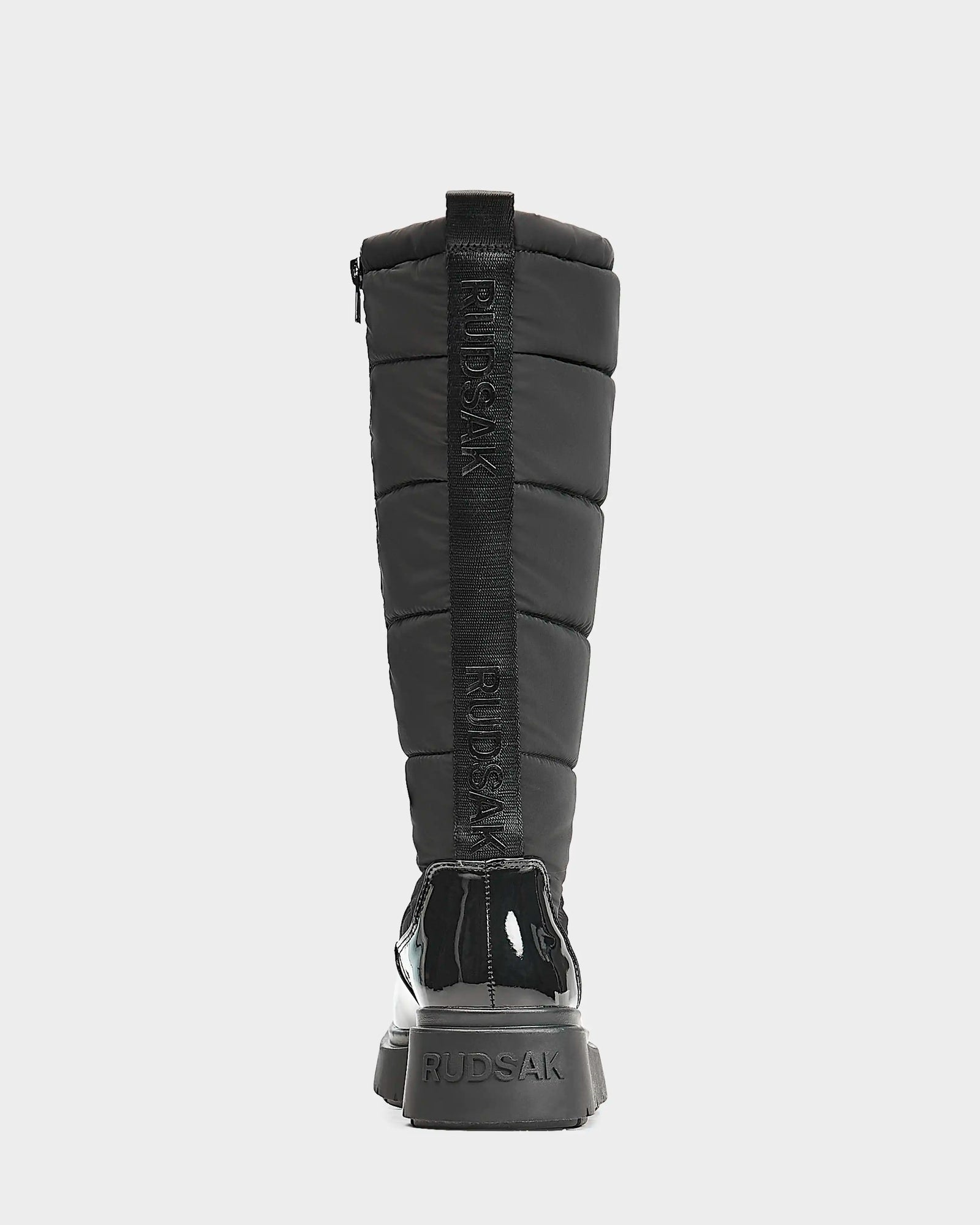 A black, knee-high women's quilted winter boot by Rudsak, CLAUDE - L223506, shown from the back with glossy heel and sole accents, featuring RUDSAK branding vertically on the back strap and heel.