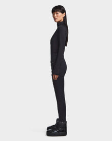 A woman stands in profile against a white background, wearing the Rudsak KILTON - 8523533 Black turtleneck, black pants, and chunky boots—perfect winter staples. Her long, straight hair falls over her shoulders.