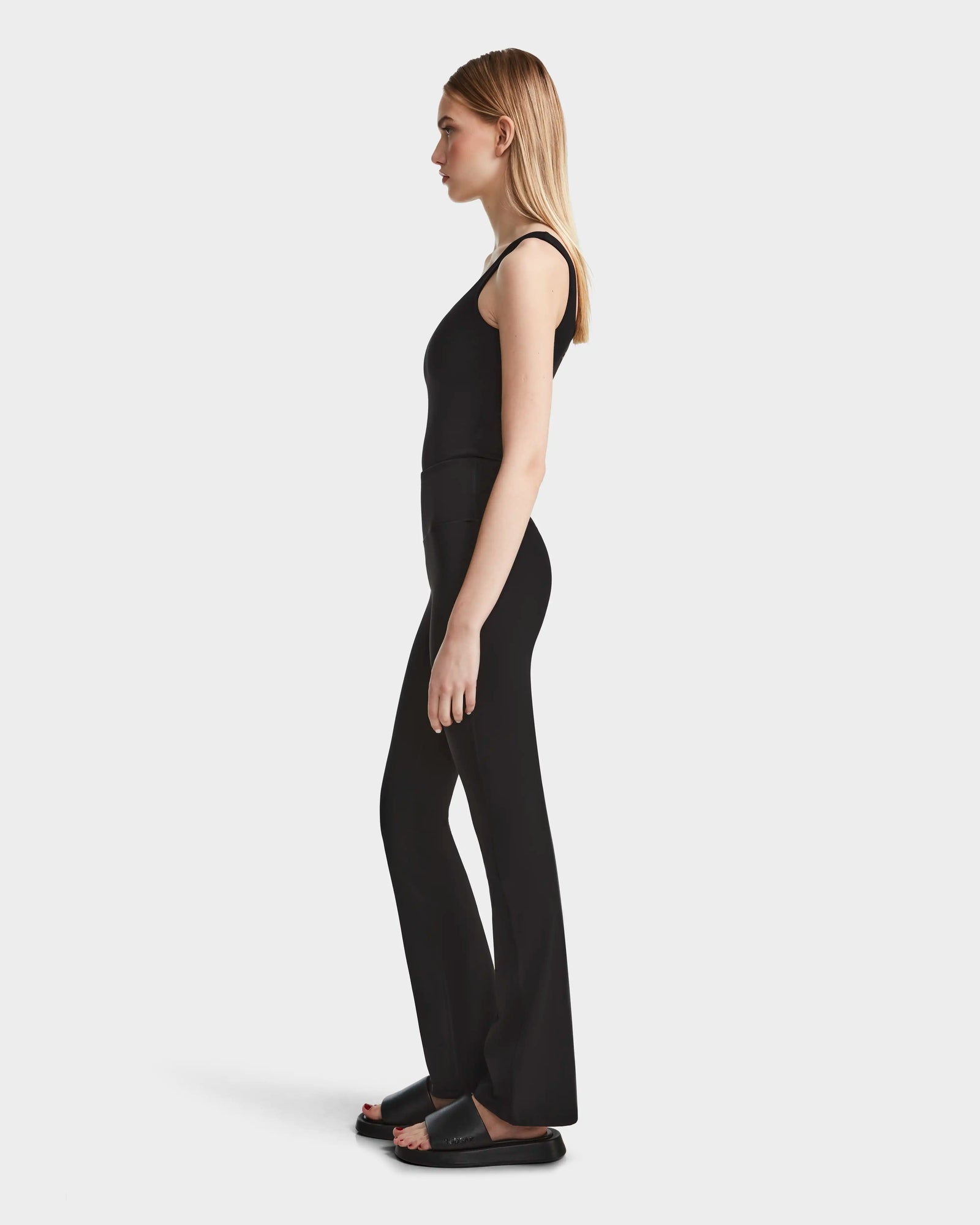 A woman with straight blonde hair stands in profile, wearing the Rudsak ADRIANA - 8524009 Black fitted sleeveless top and black flared-leg leggings, paired with black slide sandals against a plain white background.