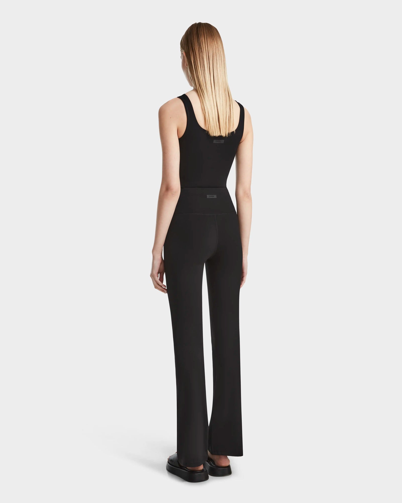 A woman with straight blonde hair stands facing away in the ADRIANA - 8524009 Black sleeveless top by Rudsak, paired with flared-leg leggings and black shoes against a plain white background.