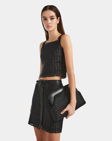 A woman with loose brown hair and a neutral expression stands against a plain background, wearing a handwoven cropped top and mini skirt, holding the Rudsak NADINE - 8524042 Black clutch—a luxury wardrobe essential.