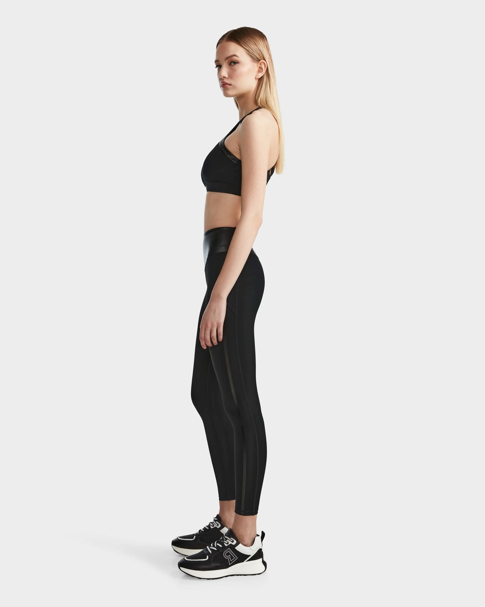 A woman with straight blonde hair poses in profile against a white background, dressed in Rudsaks THEA - 8523003 BLACK high-rise leggings with a vegan leather waistband, styled with a black sports bra and sneakers.