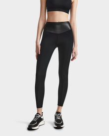 A woman models the Rudsak THEA - 8523003 BLACK high-rise leggings with a vegan leather waistband, paired with a black sports bra and sneakers, against a plain white background. Her arms are by her sides and her head is not visible.