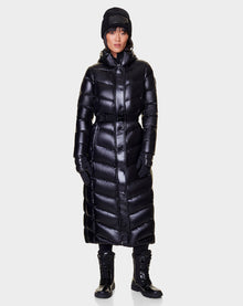 A person wearing the Rudsak ELSIE - L121525 BLACK women’s maxi down puffer, black beanie, black gloves, and black lace-up boots stands facing forward against a plain light gray background.