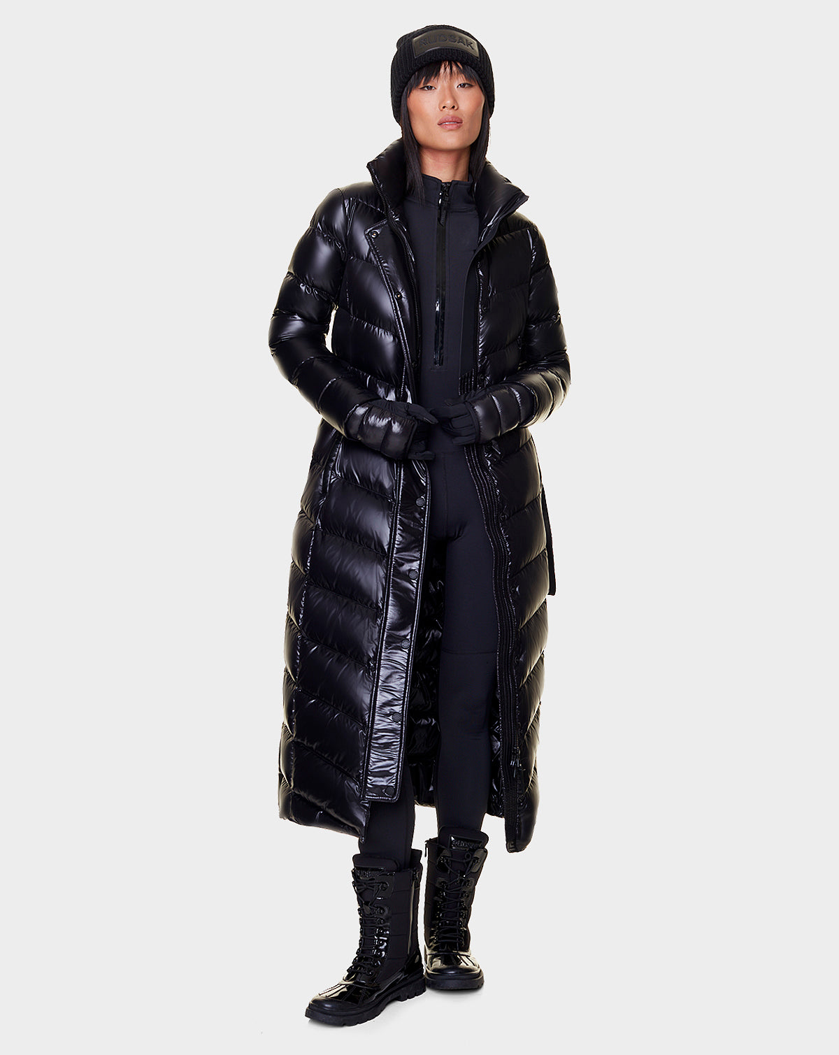 A woman wears the Rudsak ELSIE - L121525 BLACK women’s maxi down puffer with a recycled semi-gloss exterior, styled with black pants, lace-up boots, a beanie, and gloves against a plain light background.