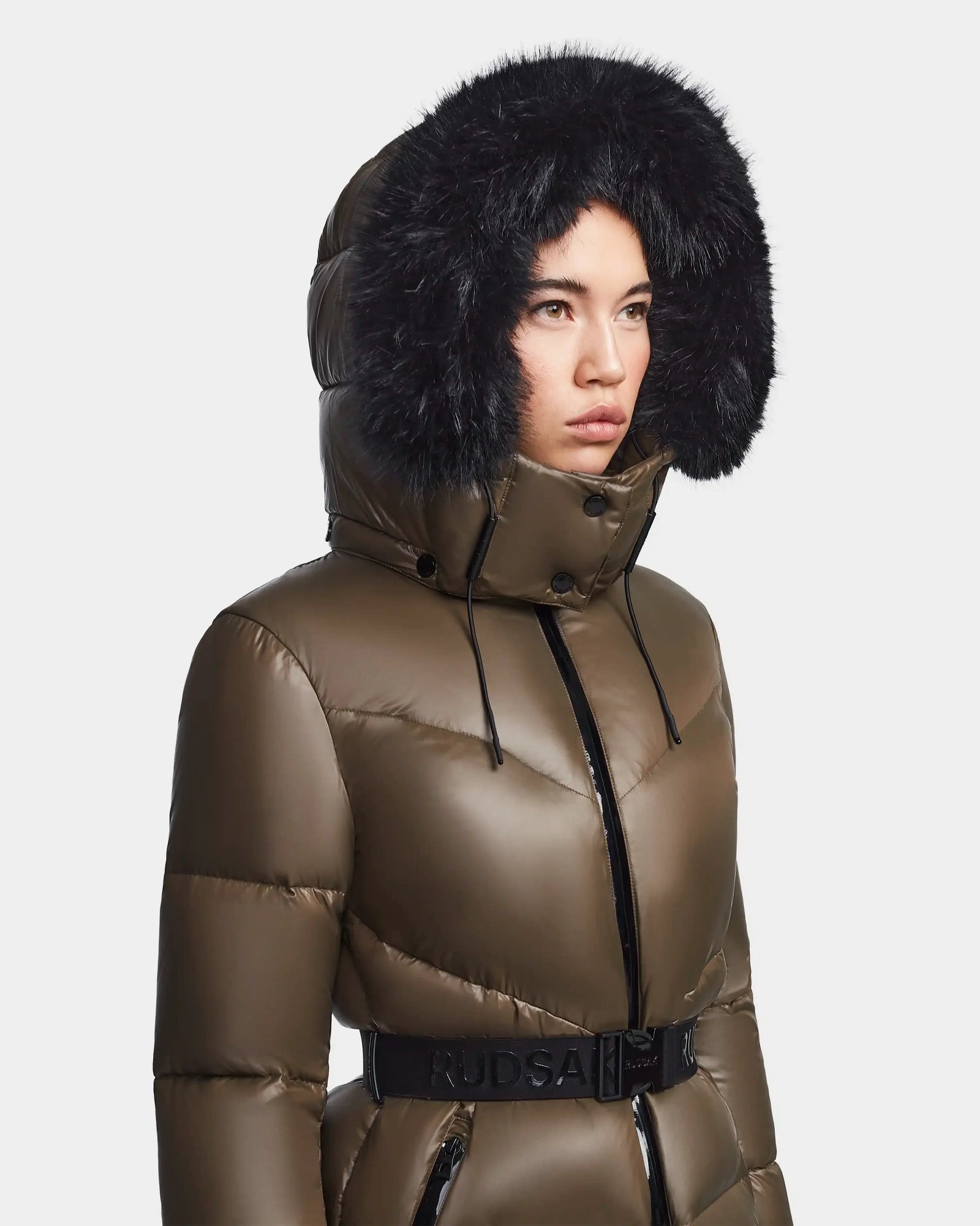 A woman wears the Rudsak CHRISTELLE S - L123524 Light Olive chevron quilted down puffer with a large black fur-trimmed hood and belted waist, looking off to the side against a plain background.