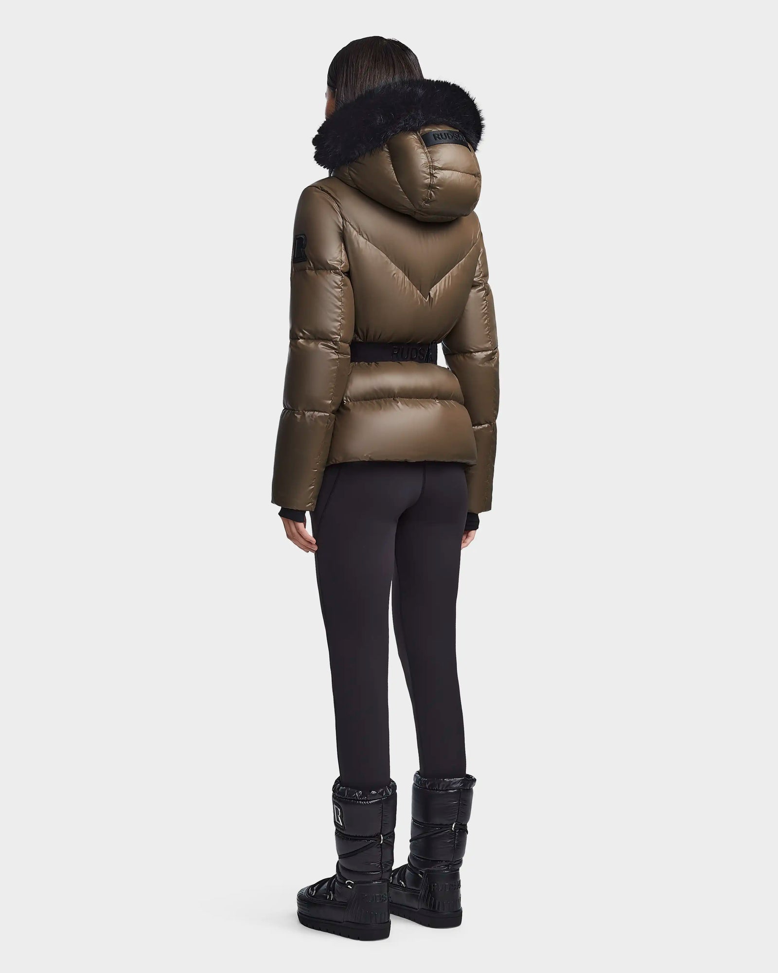 A person stands facing away, wearing the Rudsak CHRISTELLE S - L123524 Light Olive puffer jacket with a black fur-trimmed hood, paired with black leggings and winter boots against a plain light background.