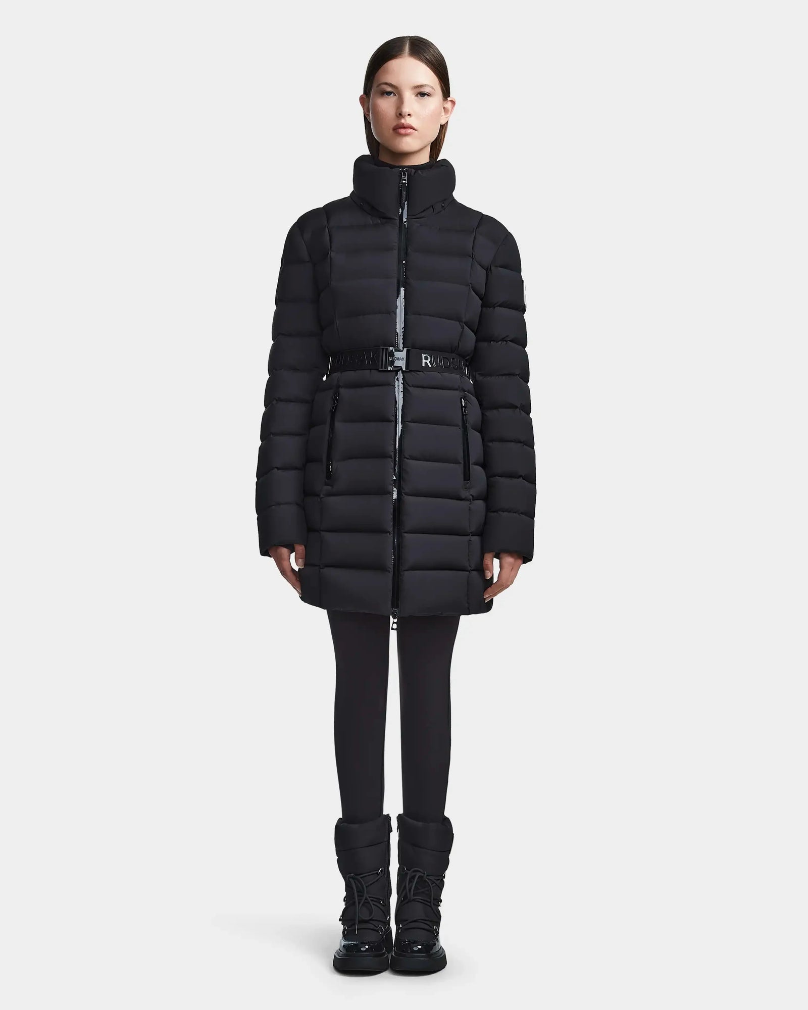 A woman faces forward, wearing the Rudsak FRIDA M - L123514 black mid-length down jacket with a removable hood, paired with black leggings and winter boots against a plain white background.