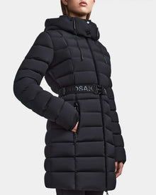Wearing the Rudsak FRIDA M - L123514 Black, a women’s mid-length down puffer coat with a belted waist, front zipper, side pockets, horizontal stitching, and removable hood. The model’s face is partially out of frame.