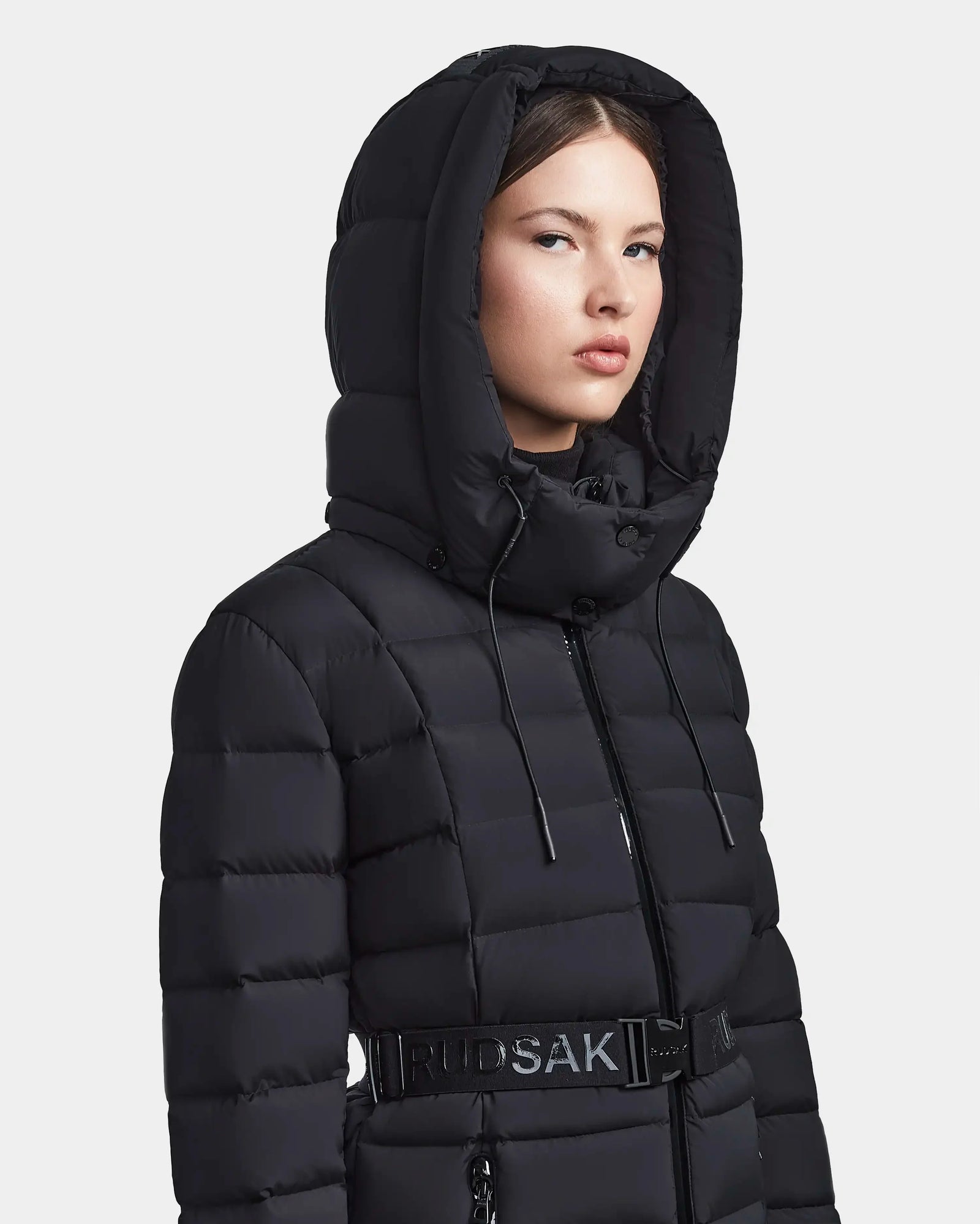 A woman wears the FRIDA M - L123514 Black, a Rudsak mid-length women’s down jacket with a large removable hood, belted waist, and Rudsak branding on the belt, facing forward against a plain background.