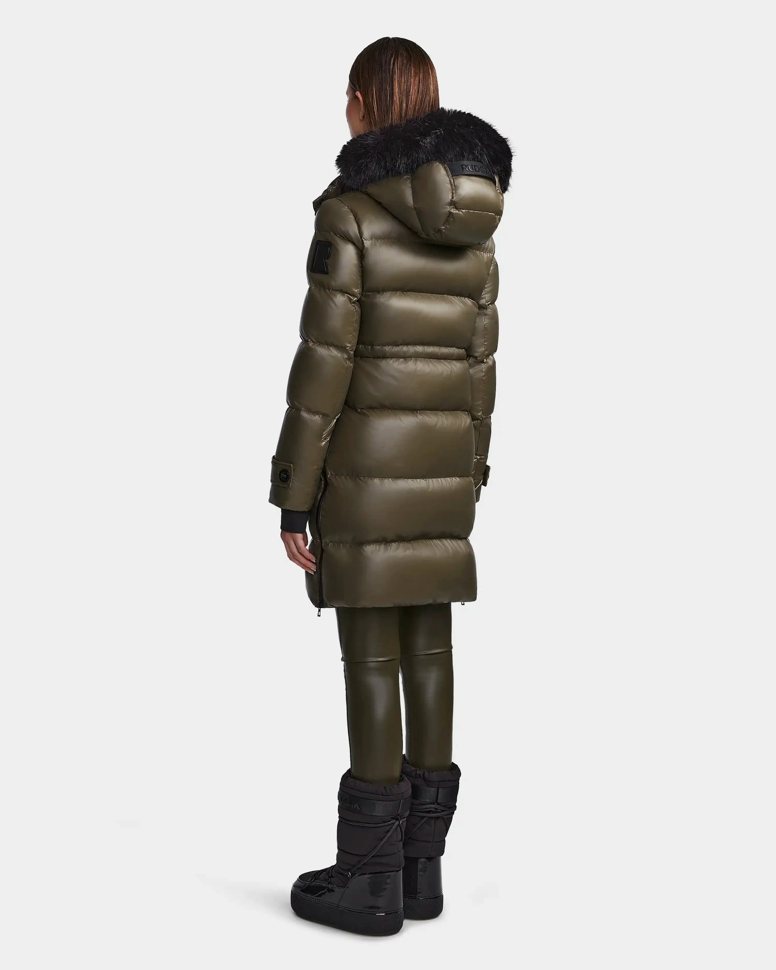 A person stands facing away, wearing the Rudsak MEYA S - L123521 Light Olive women's down puffer with a black fur-trimmed hood, matching pants, and black winter boots against a plain light background.