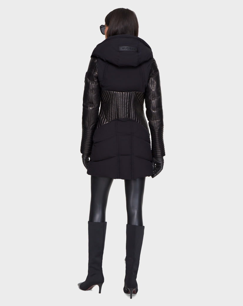 A person wearing the Rudsak SHAUNA - 8121509 BLACK quilted down puffer coat with leather sleeves, black gloves, leggings, and knee-high heeled boots stands facing away against a plain light background.