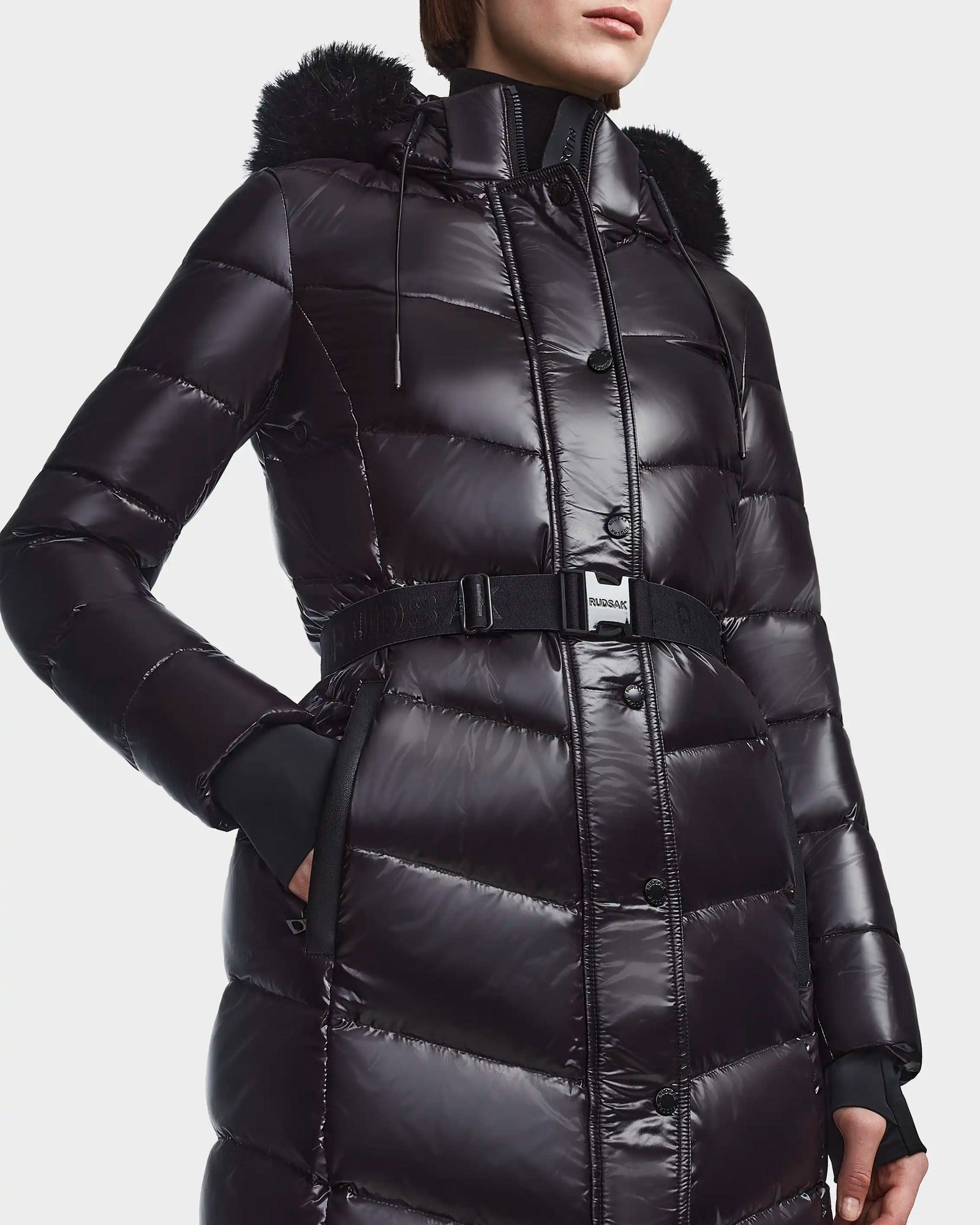 A person wears the Rudsak ELSIE S - L123518 Black puffer coat, featuring sustainable down, shiny black fabric, fur-trimmed hood, high collar, belt, horizontal quilting, and snap buttons. Only their lower face and upper body are shown.
