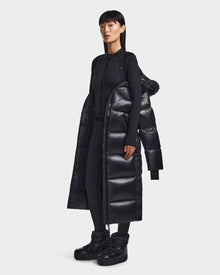 A woman with straight black hair wears the Rudsak RITA L-S - L123526 Black maxi-length down puffer draped over her shoulders, paired with a fitted black zip-up top, leggings, and platform boots against a plain white background.