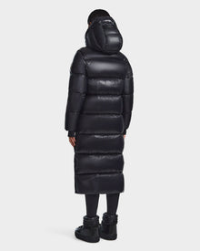 A person stands facing away, wearing the Rudsak RITA L-S - L123526 Black women's maxi-length down puffer coat with a hood, paired with black leggings and winter boots, set against a plain light background.