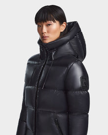 A woman with straight black hair and bangs wears the Rudsak RITA L-S - L123526 Black maxi-length down puffer coat with a hood and large sleeve 