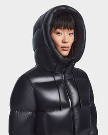 A person wears the Rudsak RITA L-S - L123526 Black puffer jacket with the hood up, featuring a shiny quilted design, padded construction, and black drawstrings, against a plain light background.