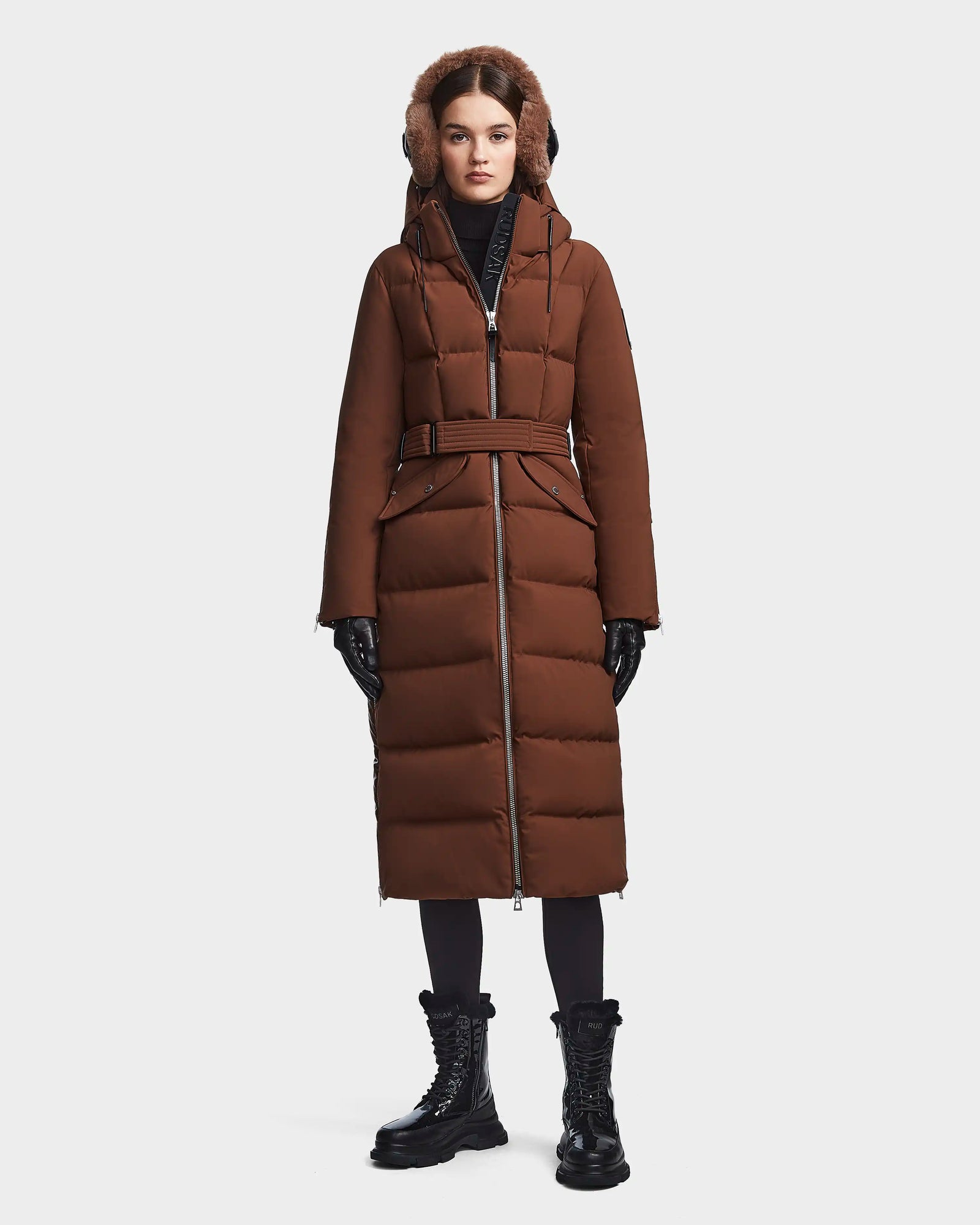A woman wears the RUDSAK CALI - 8122502 COHIBA long brown winter coat with a belted waist and fur-trimmed hood, styled with black gloves, lace-up boots, and leggings against a plain white background.