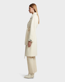 A woman stands in profile against a white background, wearing the Rudsak KOURT W - 8124702 Iceland, a long cream belted wool coat, styled with matching wide-leg pants and ankle boots.