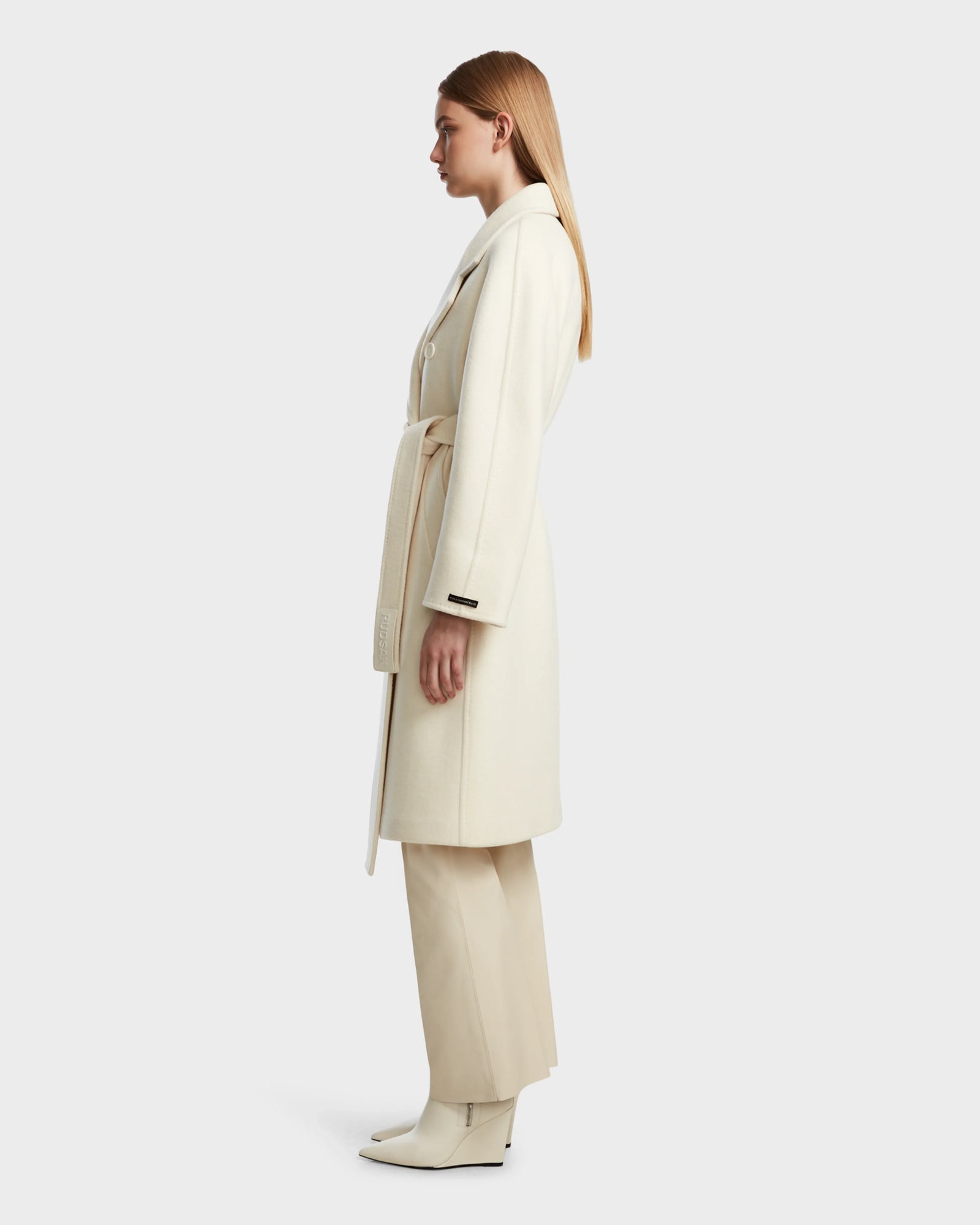 A woman stands in profile against a white background, wearing the Rudsak KOURT W - 8124702 Iceland, a long cream belted wool coat, styled with matching wide-leg pants and ankle boots.
