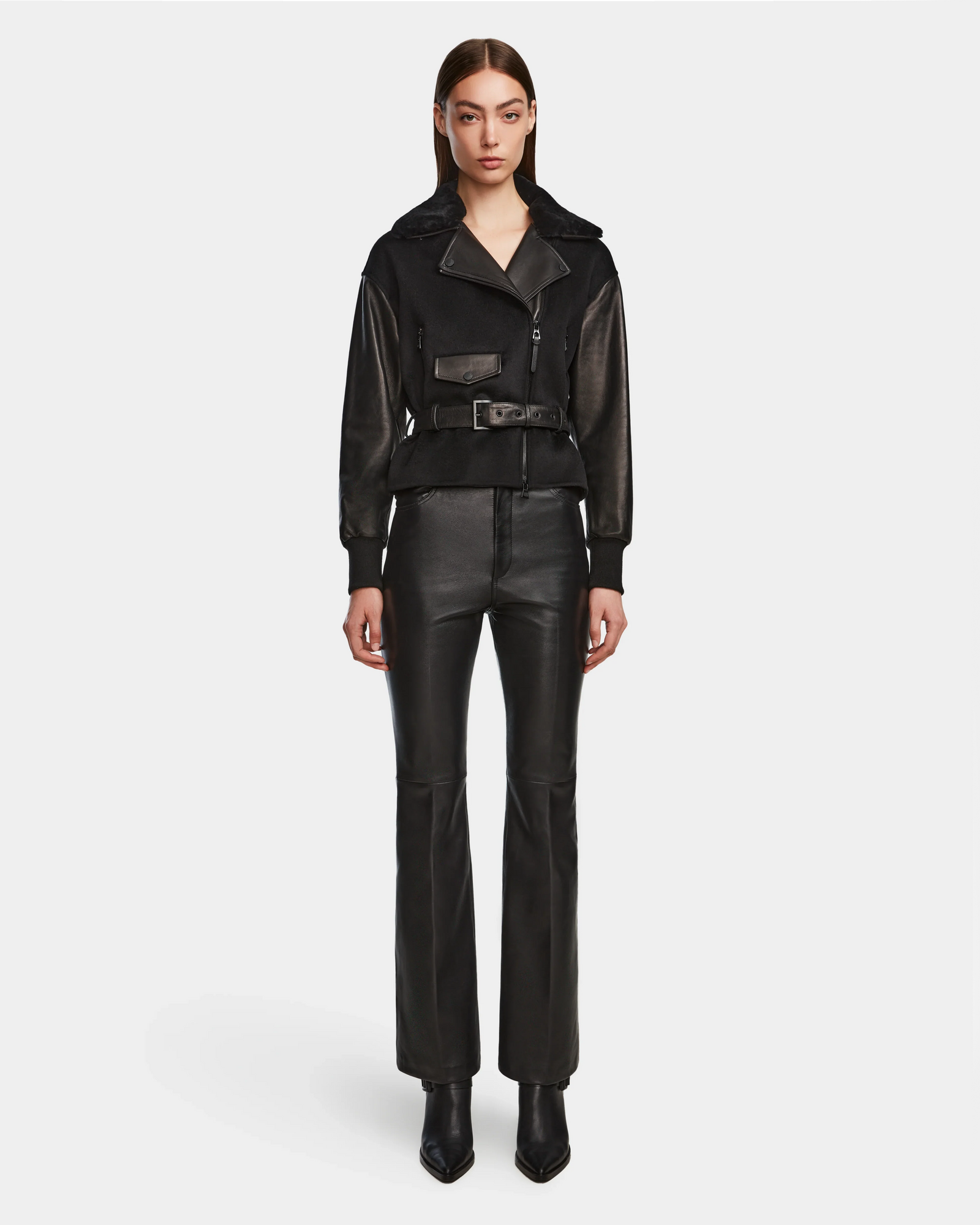 A woman faces forward in a Rudsak HALA - 8124521 Black fall jacket with a belted waist, paired with black leather pants and pointed-toe boots, set against a plain white background.